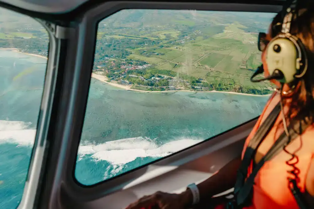 A passenger wearing a headset looks through a helicopter window towards coastal scenery, green hills and the sea.