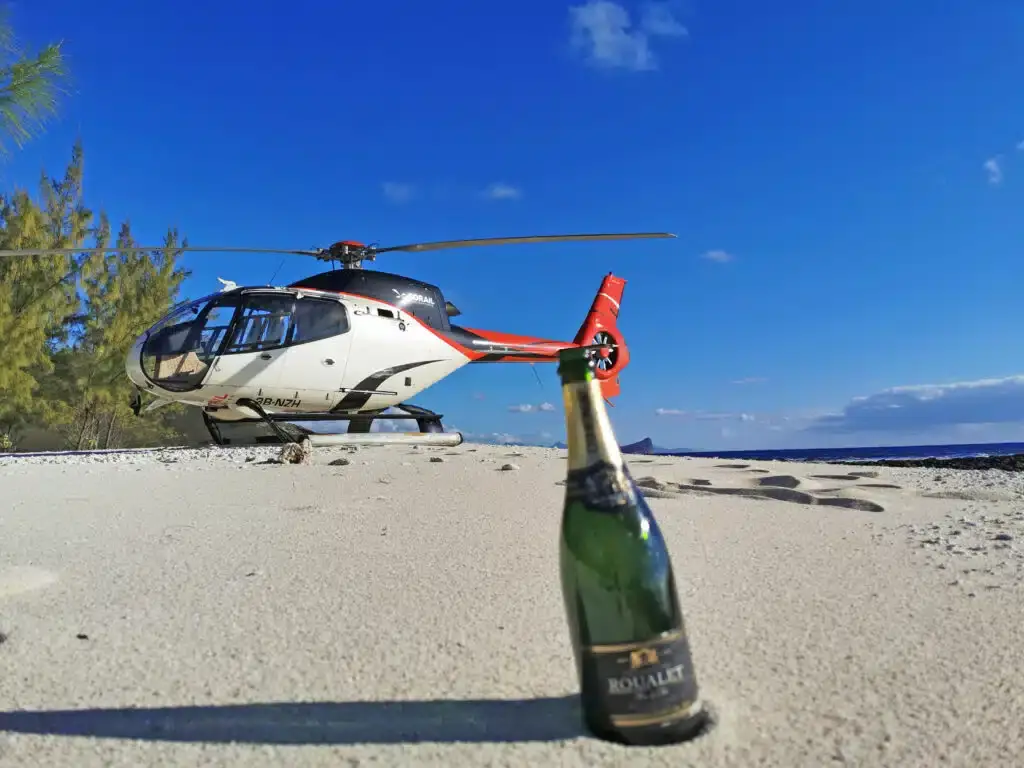 Helicopter beside a sandy beach, with a champagne bottle in the foreground and blue sky above.